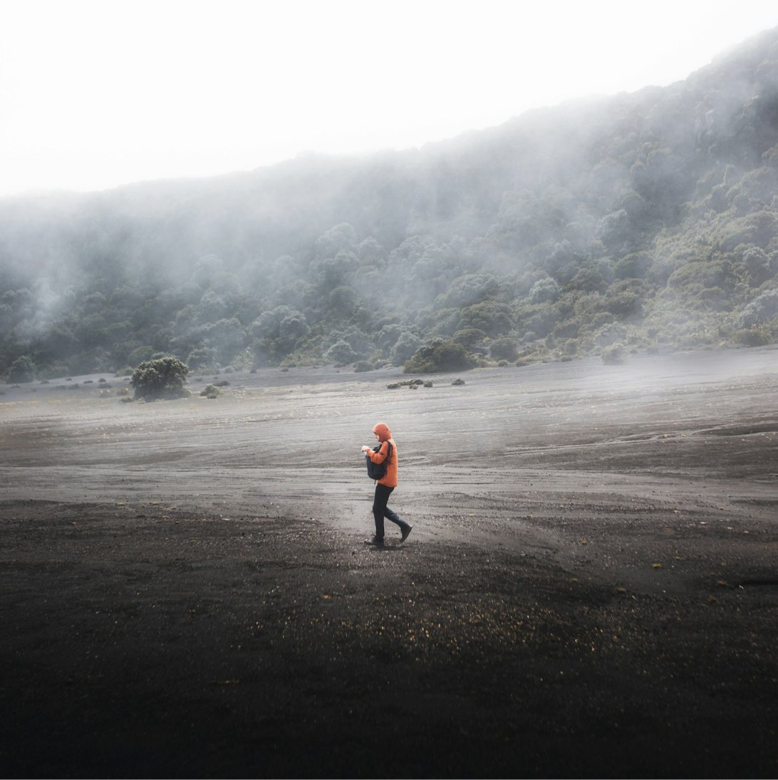 Persona con chaqueta naranja caminando en el cráter de un volcán entre la neblina